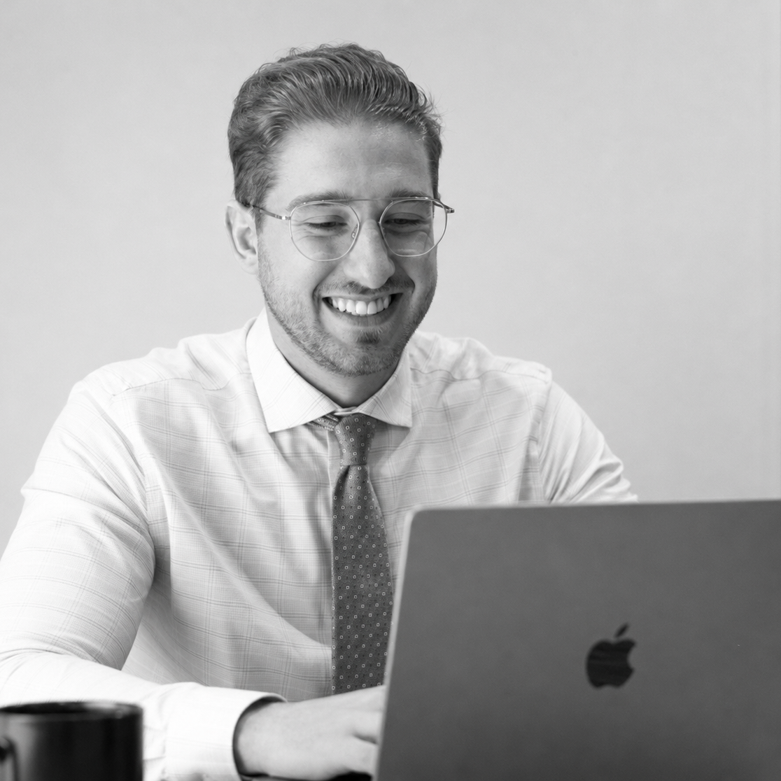 Jacob Suski - Working at desk on an advertising campaign Jacob Suski smiling with glasses working on a laptop, wearing a dress shirt and tie.